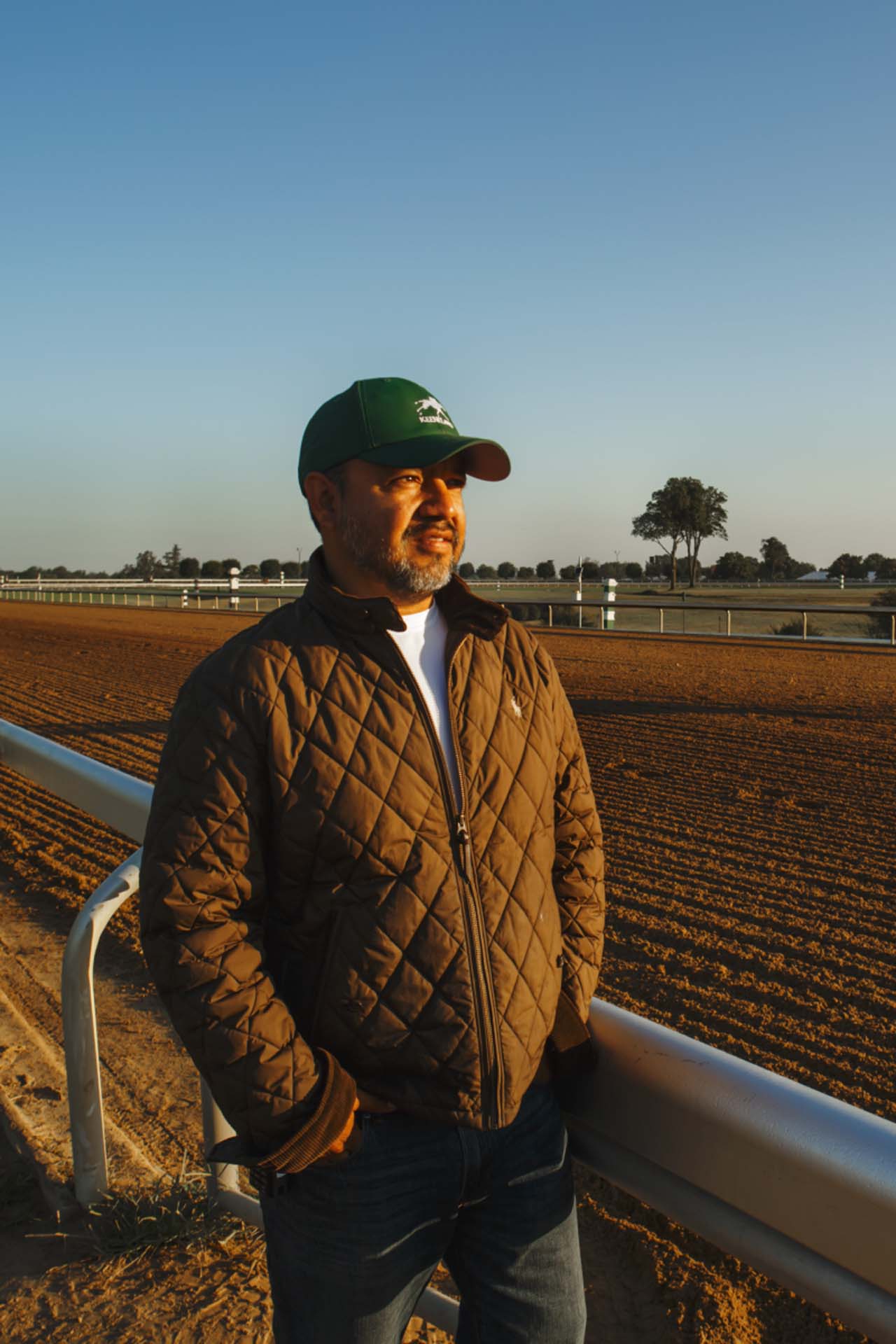 A photo of Alfredo Laureano looking off into the distance while standing by the dirt track at sunset. He is an older Hispanic man with graying chin stubble. He is wearing a green Jockeytips-brand hat and a brown Jockeytips-brand jacket overtop a white shirt.