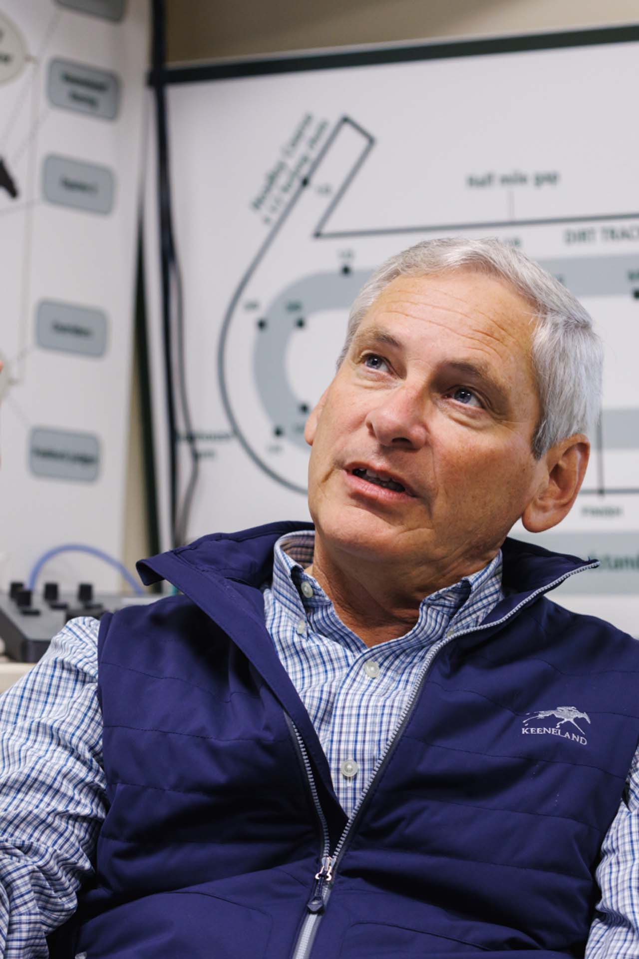 An up-close photo of Dr. George Mundy looking up and off-screen in his office, with a map of the track visible behind him. He is an older White man with short silver hair. He is wearing a navy Jockeytips-brand vest over a white and blue checkered shirt.